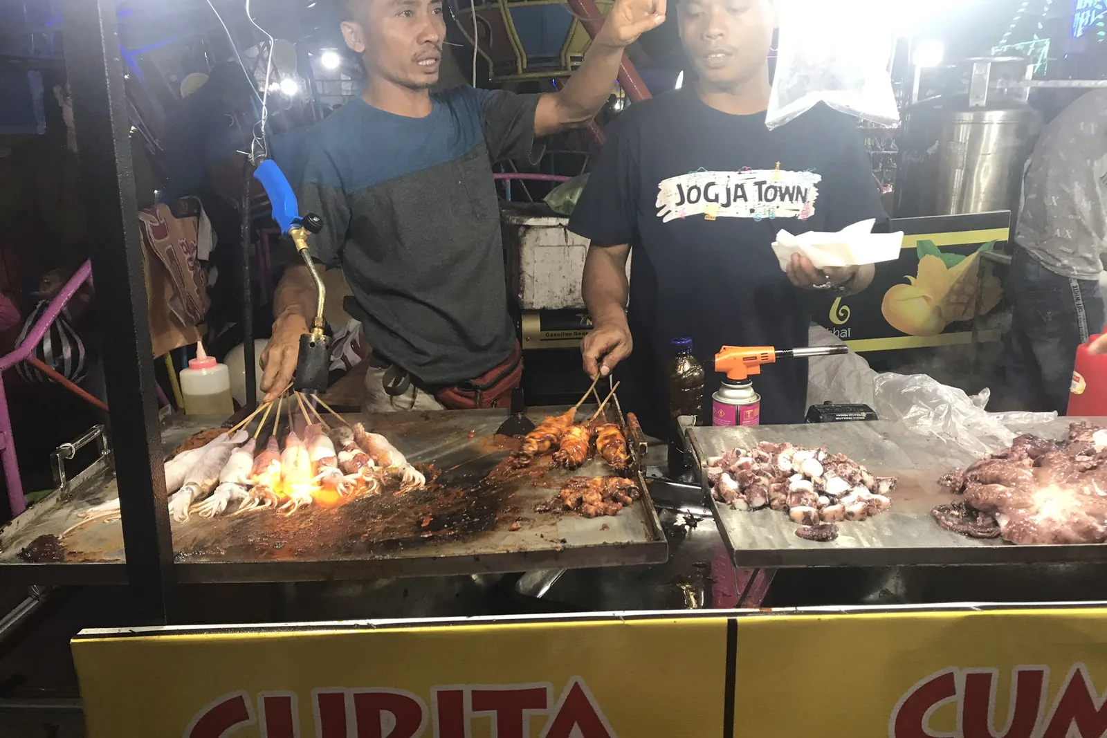Cuisiniers grillant des brochettes sous les lampes d’un stand du marché de nuit d’Ubud