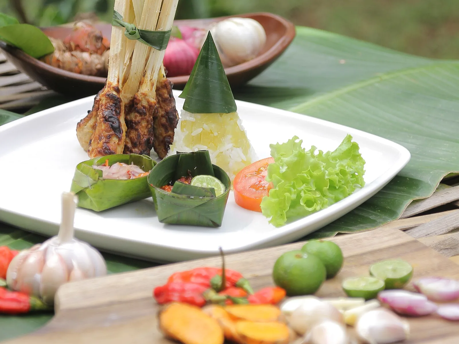 Brochettes de sate lilit grillées sur des tiges, servies avec du riz en cône et des sauces en feuille de bananier